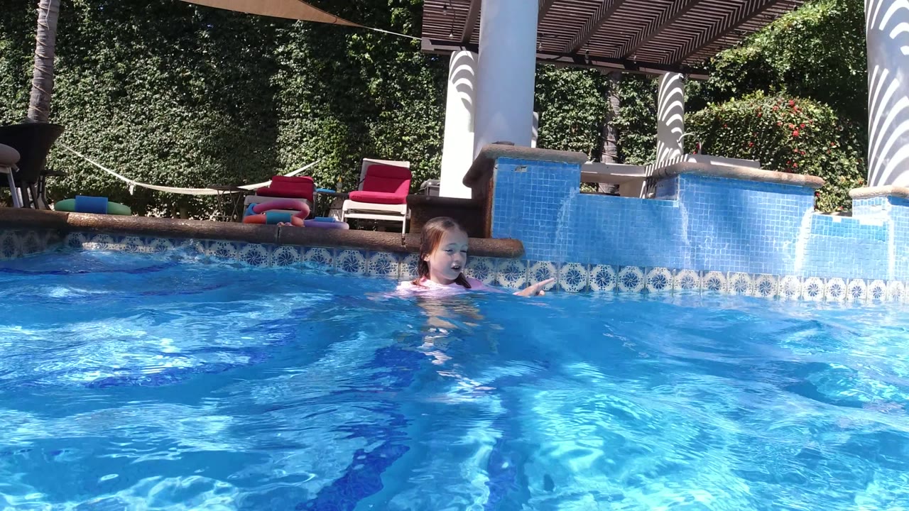 Young girl swimming in a blue pool near tiled edges and lounge chairs.