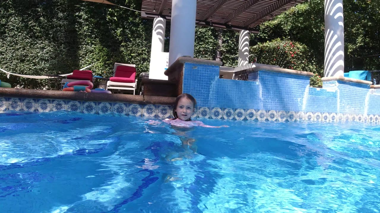At 0:01 in San Jose del Cabo, Mexico, the girl is closer to center frame in the pool, with sunlit blue water in front and shaded lounge chairs and dense greenery in the background.