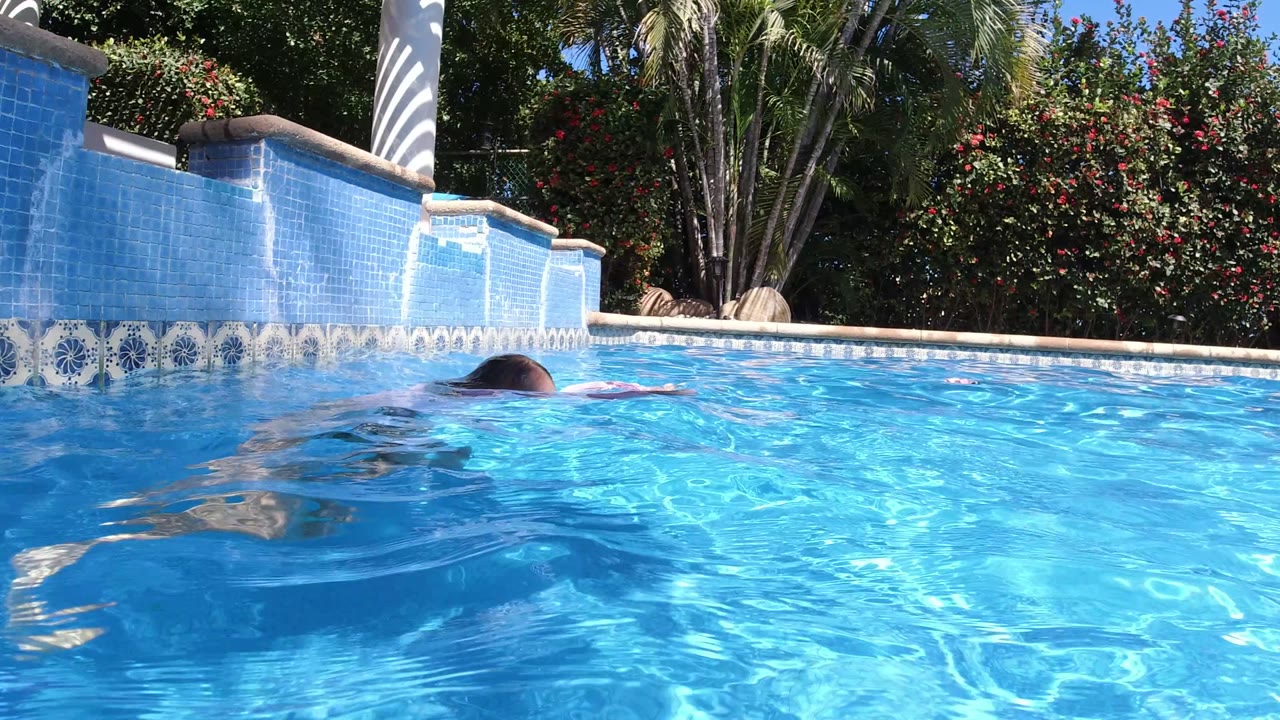 The child surfaces farther across the San Jose del Cabo pool, with rippled blue water in front and the garden wall and palms in the background.