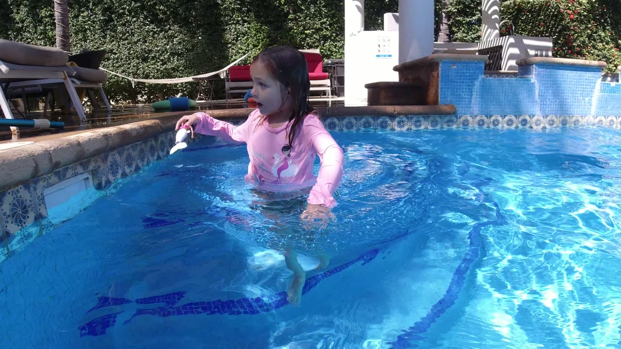 Blonde child in a pink swim shirt standing in a blue pool with a white toy.