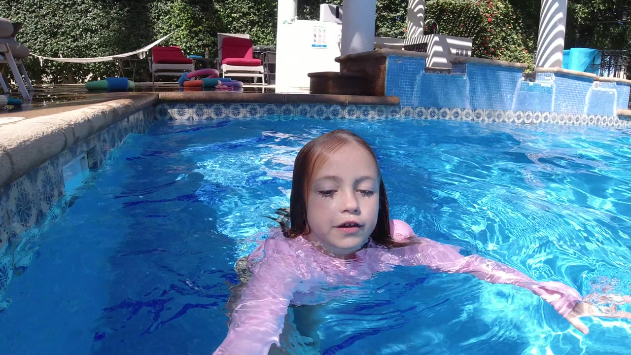 Young girl in a pink long-sleeve swim dress floats near the camera in bright blue pool water in San Jose del Cabo, Mexico, with patio furniture and hedges behind her.