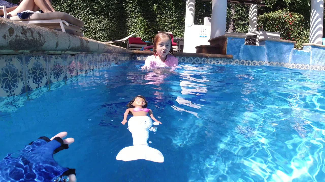 At 0:00 in San Jose del Cabo, a red-haired girl faces the camera from the pool while a mermaid doll floats in the foreground and an adult lounges by the pool edge.