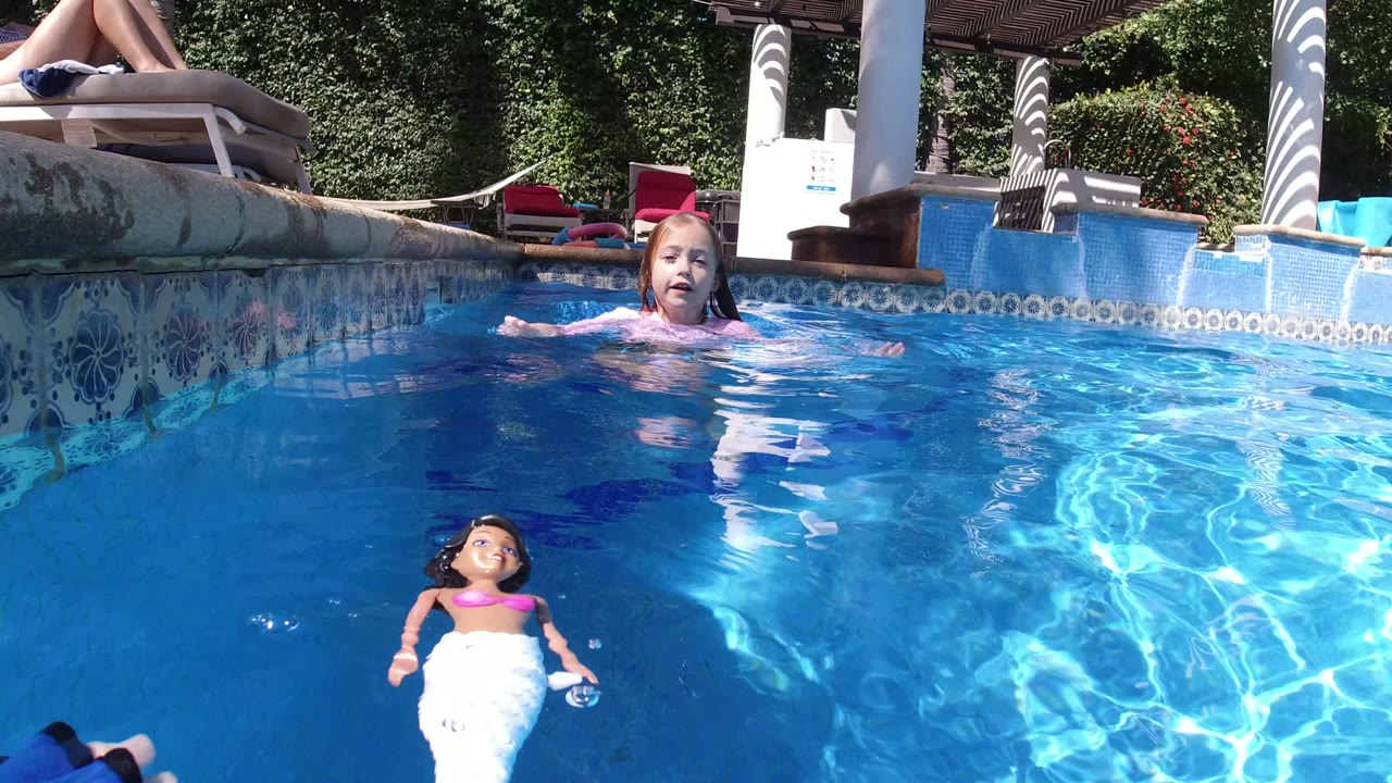 At 0:01 in San Jose del Cabo, the girl remains near the far side of the pool while the mermaid doll floats lower in frame amid rippled blue water.