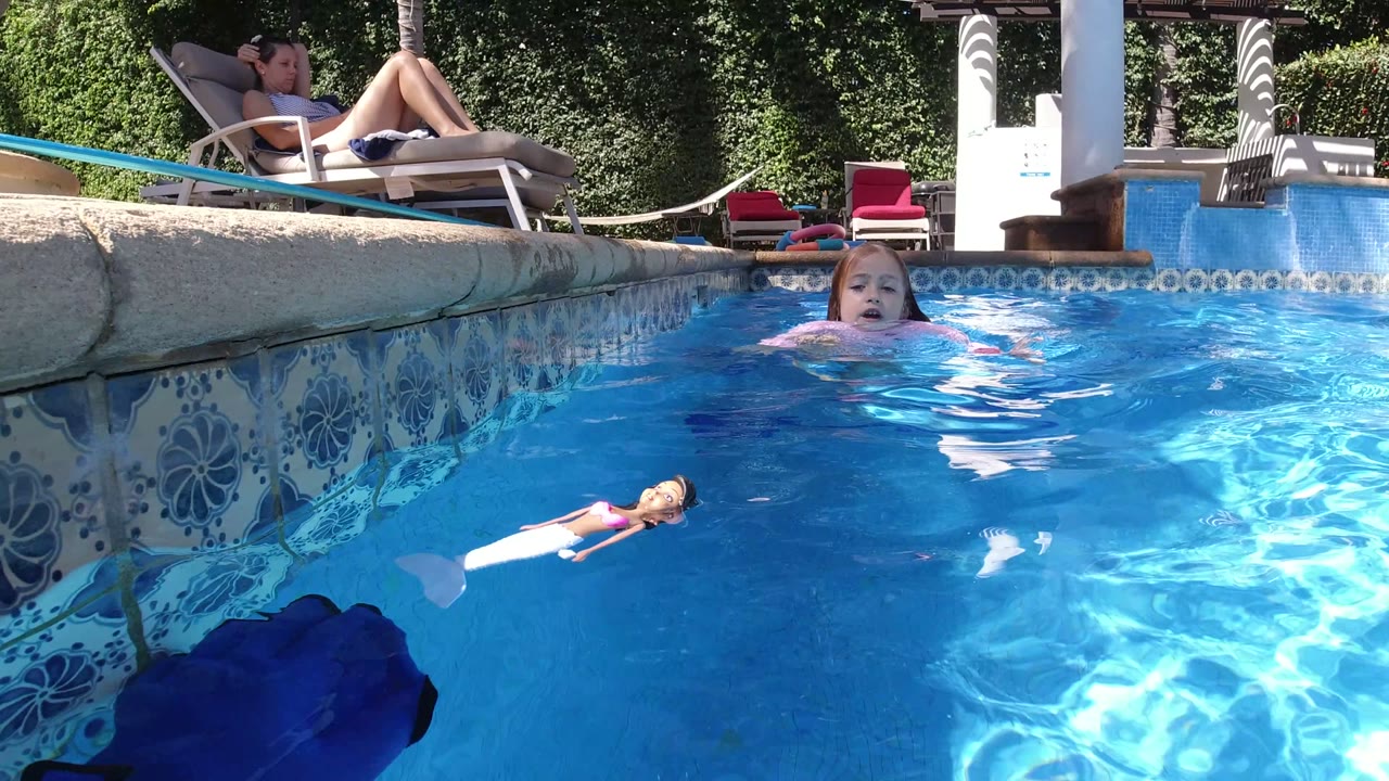 At 0:00 in San Jose Del Cabo, Mexico, a young girl faces the camera in a sunlit pool while a doll floats in front of her and an adult reclines on a lounge chair behind the pool wall.