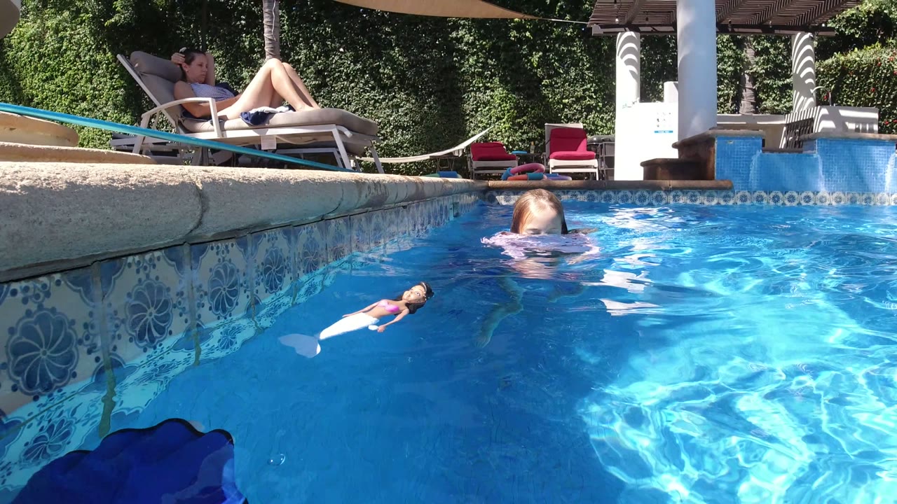 At 0:01 in San Jose Del Cabo, Mexico, the girl is partly turned away in the blue pool, with the doll floating closer to the foreground and lounge seating visible above the pool edge.