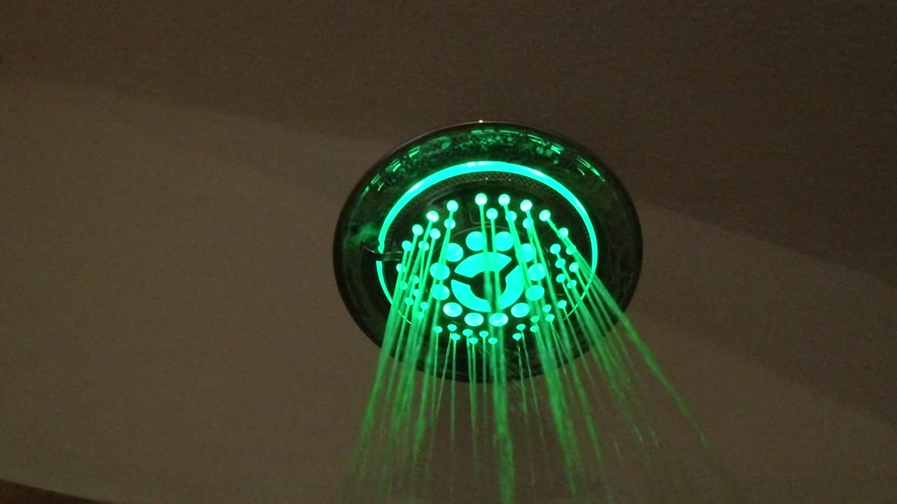 At 0:01, the shower head remains centered high in frame as green light catches the falling water, with no visible people.