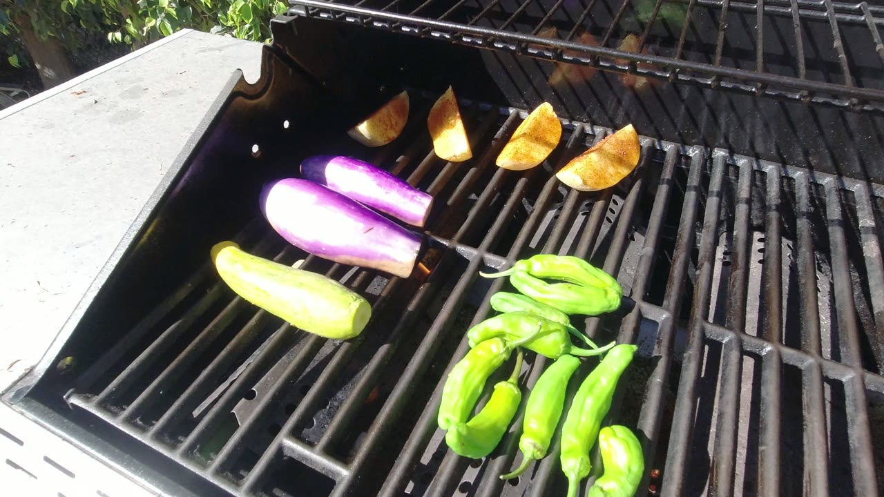 Sunlit Los Cabos barbecue frame with green peppers clustered at lower right, purple eggplant near center, squash at left, potato wedges along the back, and no visible people.