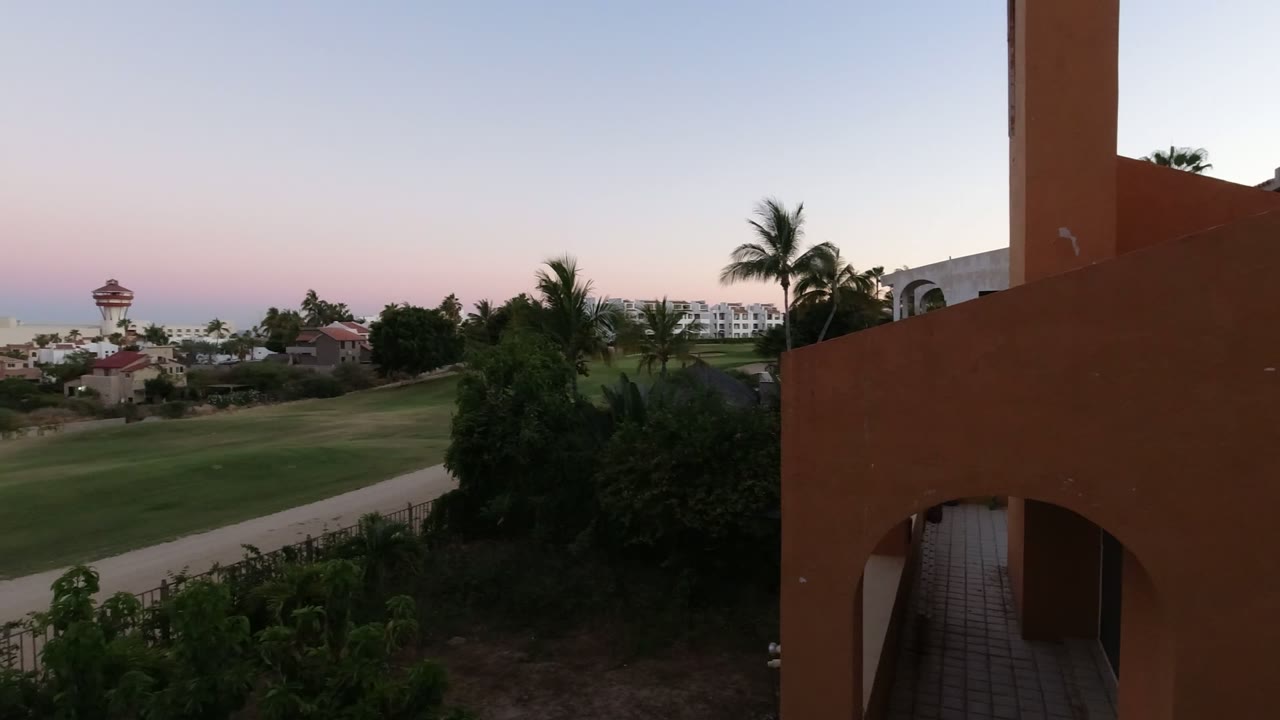 At 0:01 in Los Cabos, the elevated view keeps the orange wall and arch at right while showing the lawn, palms, distant buildings, and pink-blue dusk sky with no visible people.