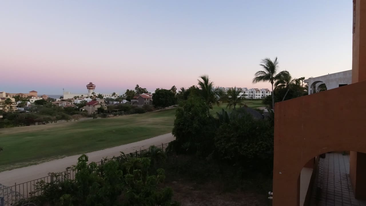 Sunset view over a green fairway, palms, buildings, and distant ocean from a balcony.
