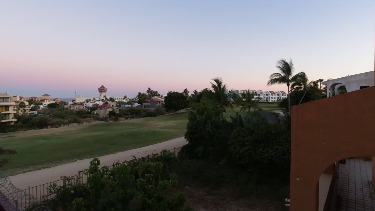 Los Cabos sunset frame with the fairway and path centered, palms on the right, distant buildings along the horizon, ocean beyond them, and no visible people.