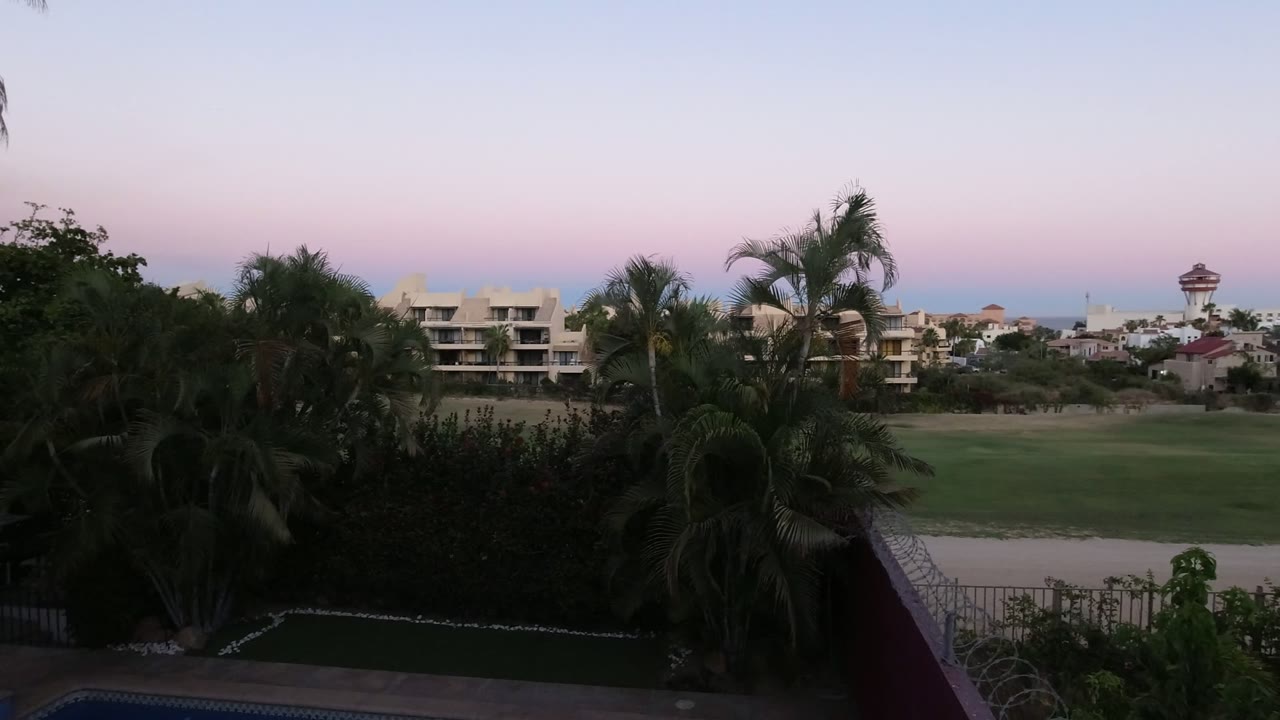 Opening frame of a Los Cabos balcony view with dark palms in front, pale buildings beyond the lawn, a thin ocean line, and no visible people.