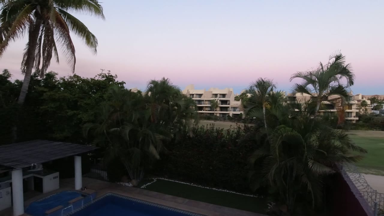 High view over a pool, palms, hedges, and pale buildings at dusk.