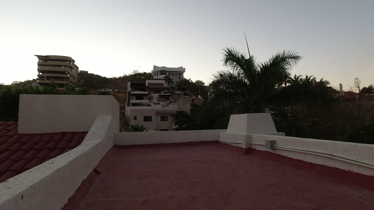 Red rooftop terrace with white parapet, hillside homes, trees, and palms at dusk.
