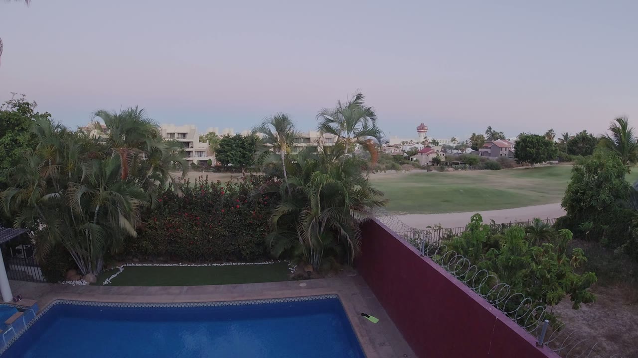 Second frame shows the Los Cabos pool view held from above, with the red wall more central, palms filling the left and middle, open green grounds beyond, and no visible people.