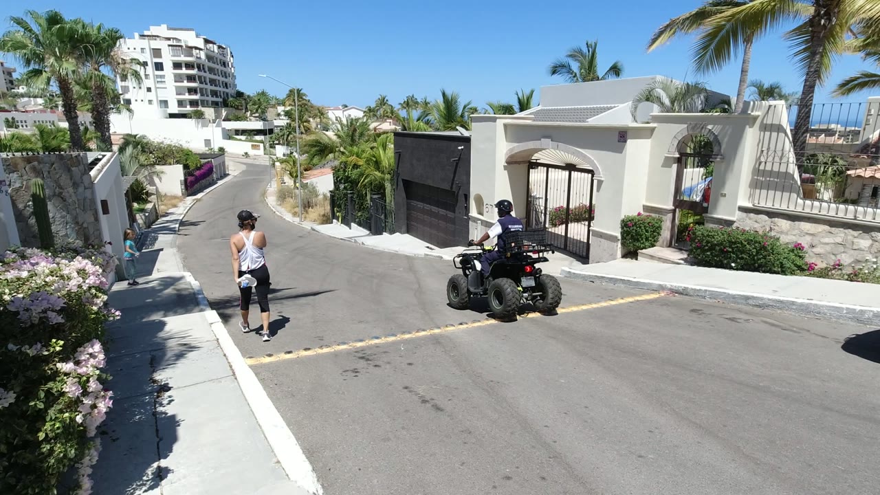 A woman walks along the Los Cabos street while an ATV rider moves past a white gated property, with palms, stone walls, and a hillside building behind them.