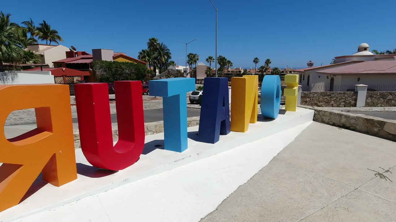 Opening view of colorful freestanding letters on a white concrete foundation in Los Cabos, Mexico, with palms, walls, houses, blue sky, and no visible people.