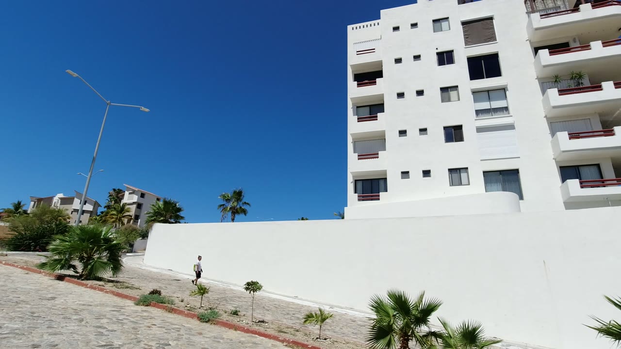 The man is slightly farther along the Los Cabos sidewalk, with the white wall filling the midground and the apartment-style building cropped near the upper right.