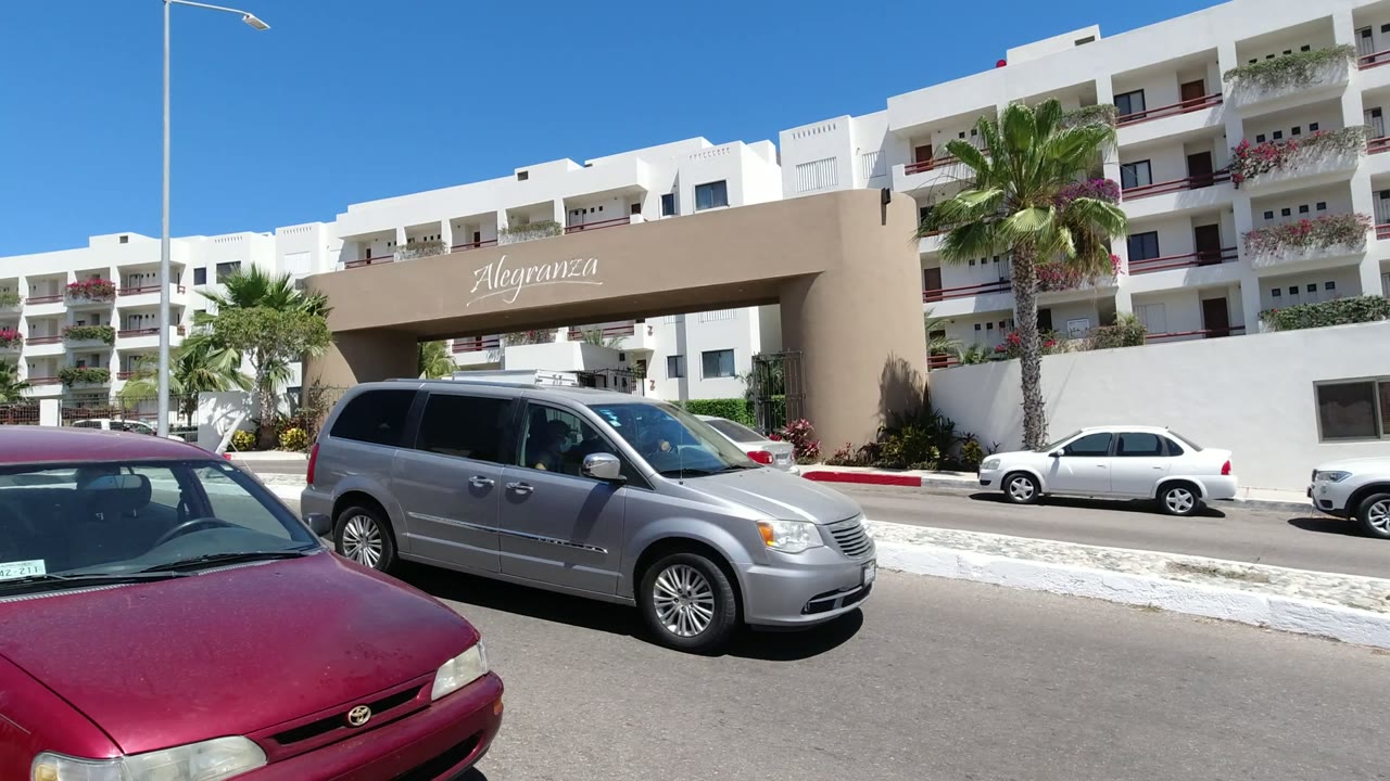 A gray minivan moves through the foreground on a Los Cabos street while the Alegranza entrance, white buildings, palms, and no visible people sit behind it.
