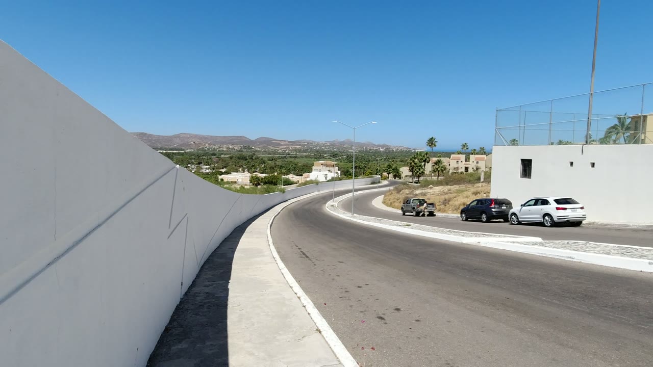 Curved road beside white walls with parked vehicles and distant hills.