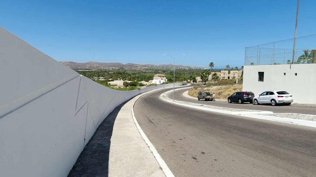 Los Cabos street frame with the sidewalk leading toward a bend, white stucco-like walls, a few parked cars, low homes, palms, hills, and no visible people.