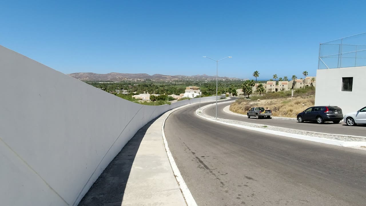Curving road bordered by white walls with cars and distant hills.