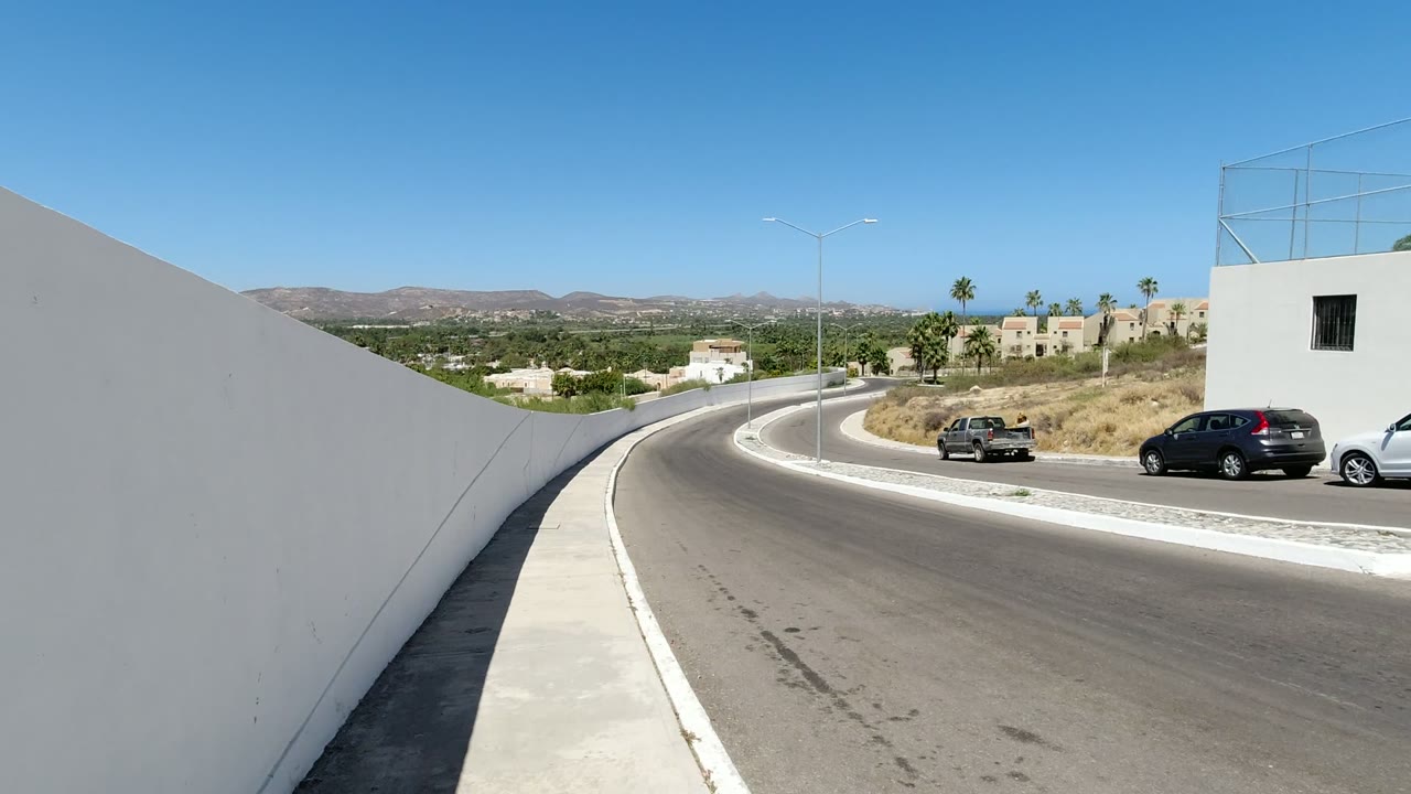 Second frame shows the same Los Cabos road from a high roadside angle, with cars spaced along the curve, white walls in the foreground, dry hills beyond, and no visible people.