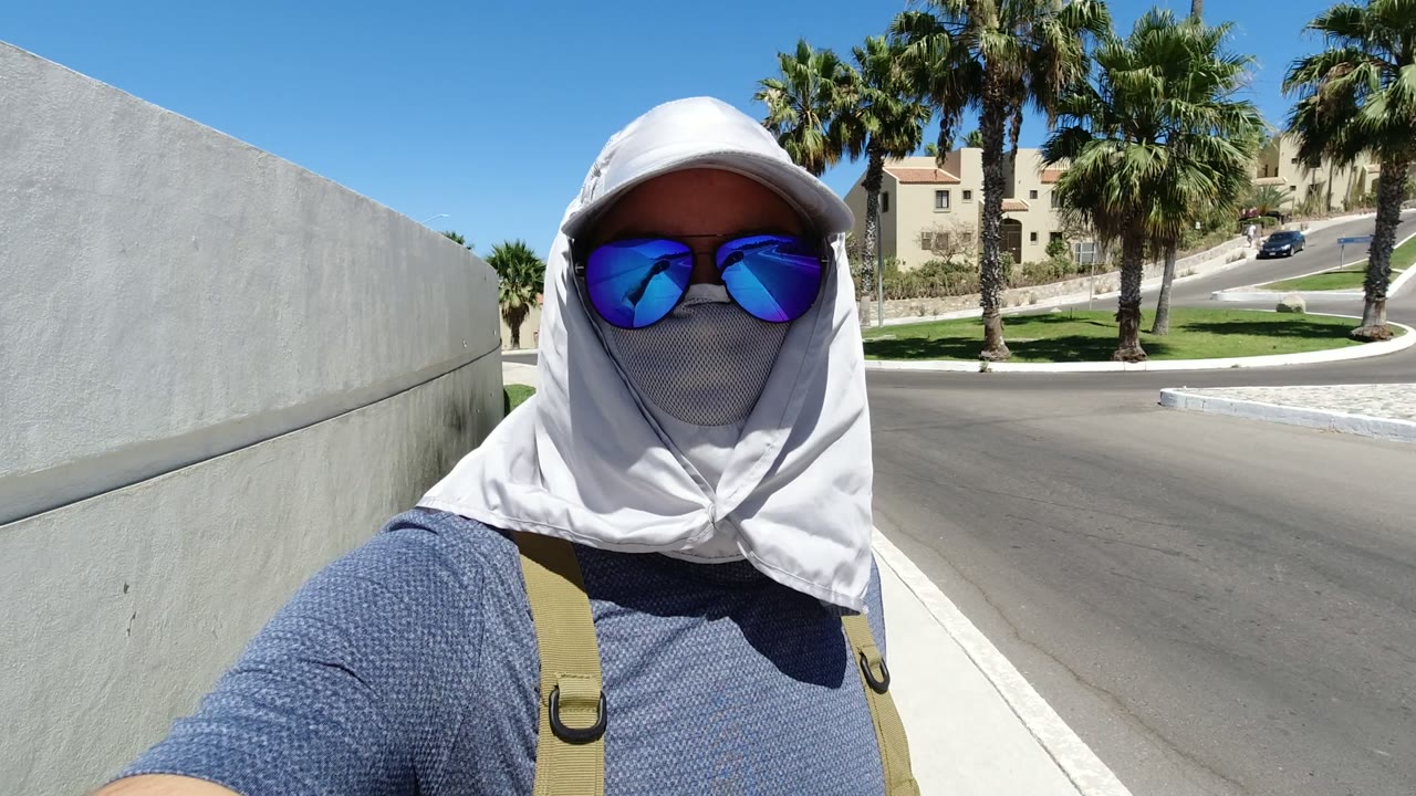 At 0:01 in Los Cabos, the person remains close to camera with the pale wall extending behind them and palm trees, road, and low buildings visible in strong sun.