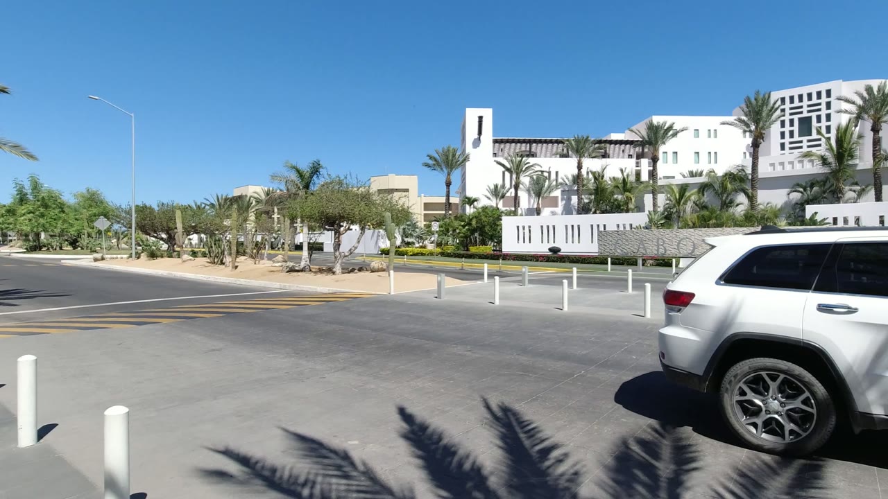 The white SUV moves farther across the foreground in Los Cabos, Mexico, partially blocking the signed entrance wall while palm trees and white buildings remain visible behind it.