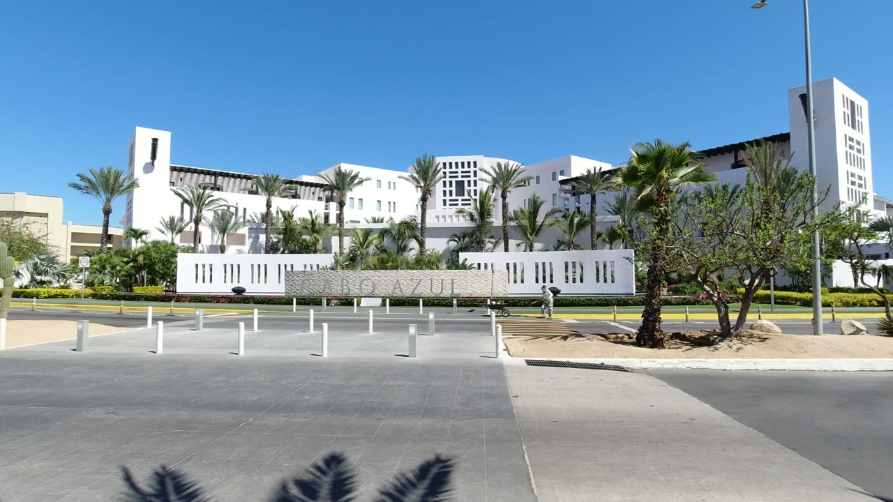Second frame holds on the Los Cabos resort wall and palms, with a taller white building section at right, road in front, and no clearly visible people.