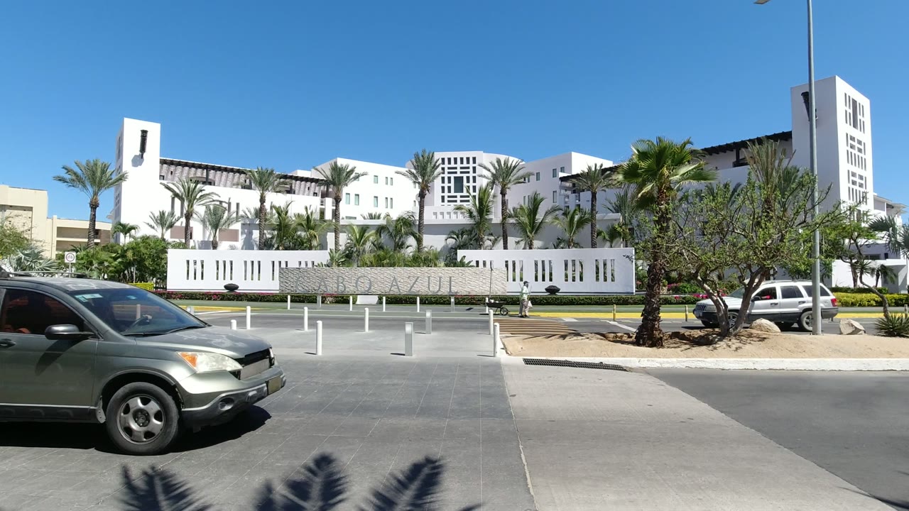 Opening Los Cabos frame with a gray SUV partly visible at left, white bollards in the foreground, palms, and a large white modern building under clear blue sky, no visible people.