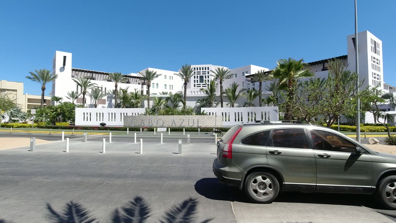 Los Cabos street view one second in, with the road open across the foreground, white posts, palm landscaping, and the modern white building set behind a signed wall, no visible people.