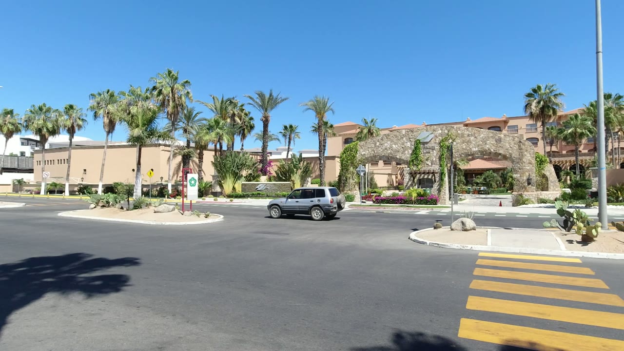 A silver SUV moves across the Los Cabos street while the yellow crosswalk, landscaped island, palms, and tan buildings remain in clear sun with no visible people.