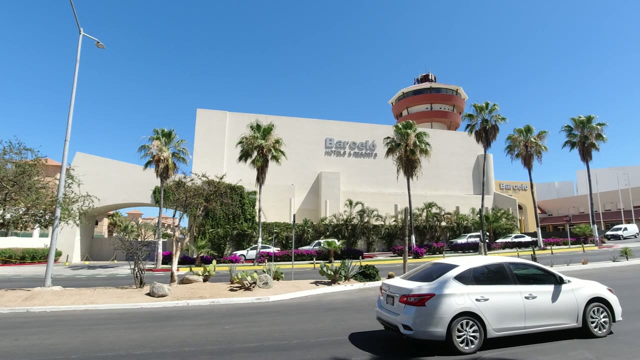 At 0:01 in Los Cabos Mexico, the mall entrance area and round tower sit behind palms and desert landscaping while a white sedan crosses the foreground road, with no clearly visible people.