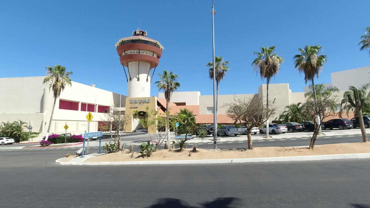 White building with round tower, palms, parked cars, and road foreground.