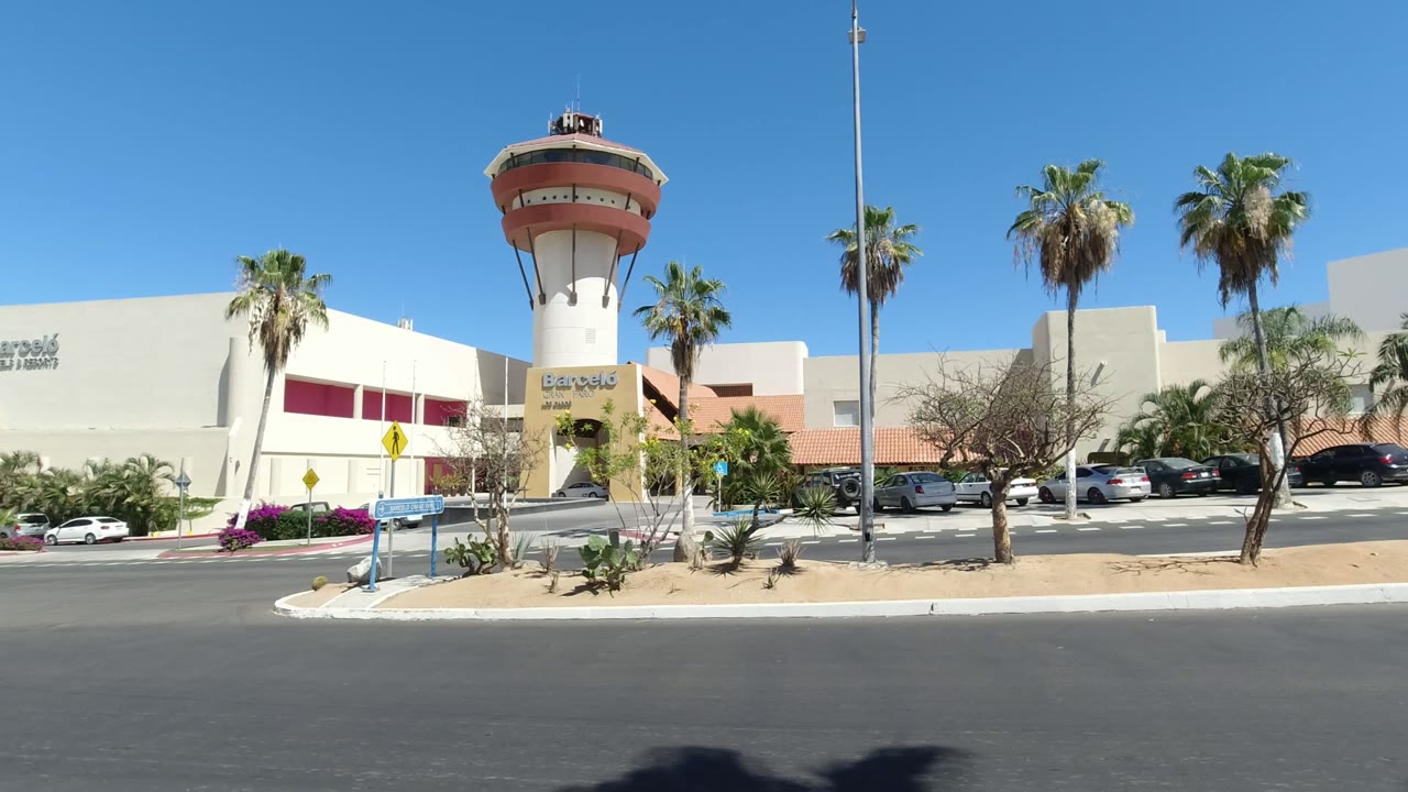 Second frame of the Los Cabos Mexico exterior, with the round tower slightly more centered, palm trees on both sides, a white facade, parked cars, visible signage, and no visible people.