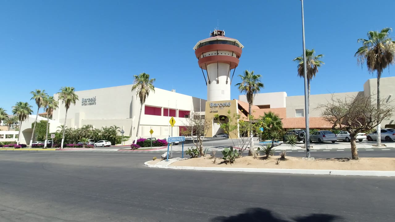 Wide view of a white and red shopping plaza in Los Cabos, Mexico, with a tower entrance, palm trees, parked cars, road foreground, and no clearly visible people.