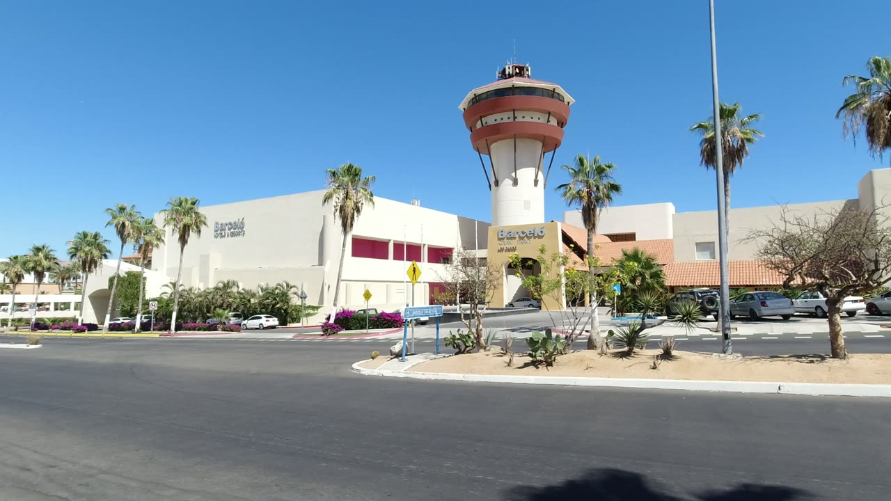 The Los Cabos plaza remains centered with the tower-like entrance, red facade panels, palm trunks, parked cars, landscaping, and no clearly visible people.
