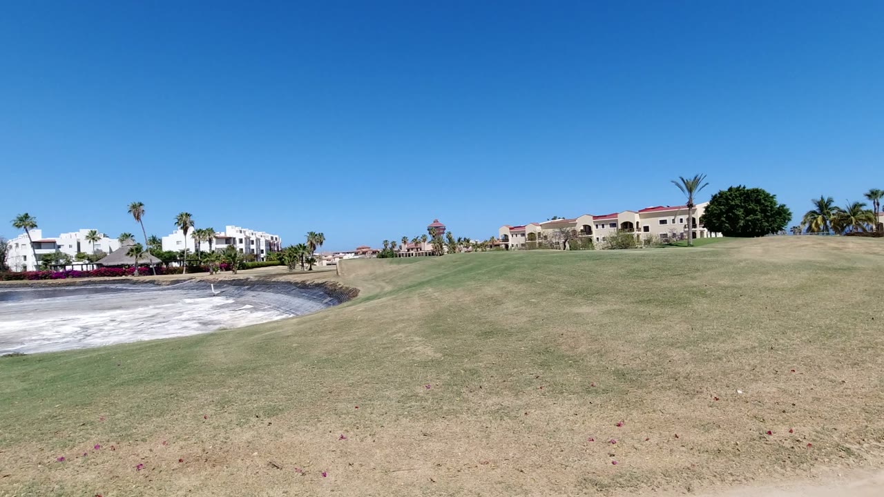 Wide lawn beside a pale circular pit and resort buildings under blue sky.