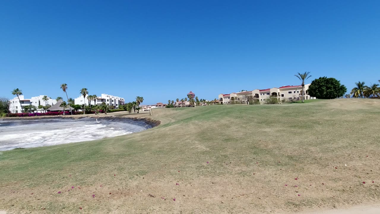 Los Cabos resort grounds with a broad grass slope leading toward a pale circular pit, white buildings, palm trees, and no visible people.