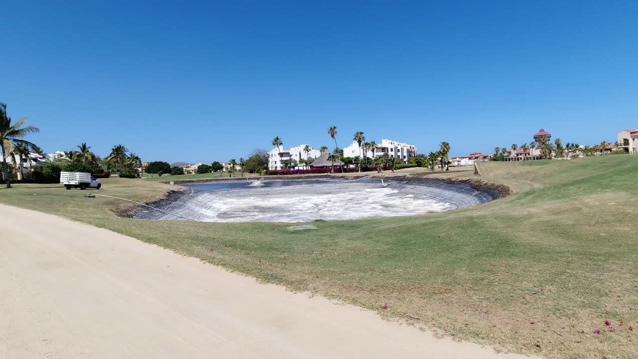 Wide Los Cabos exterior with a sandy path in the foreground, clipped grass, palm trees, distant buildings, and no visible people.