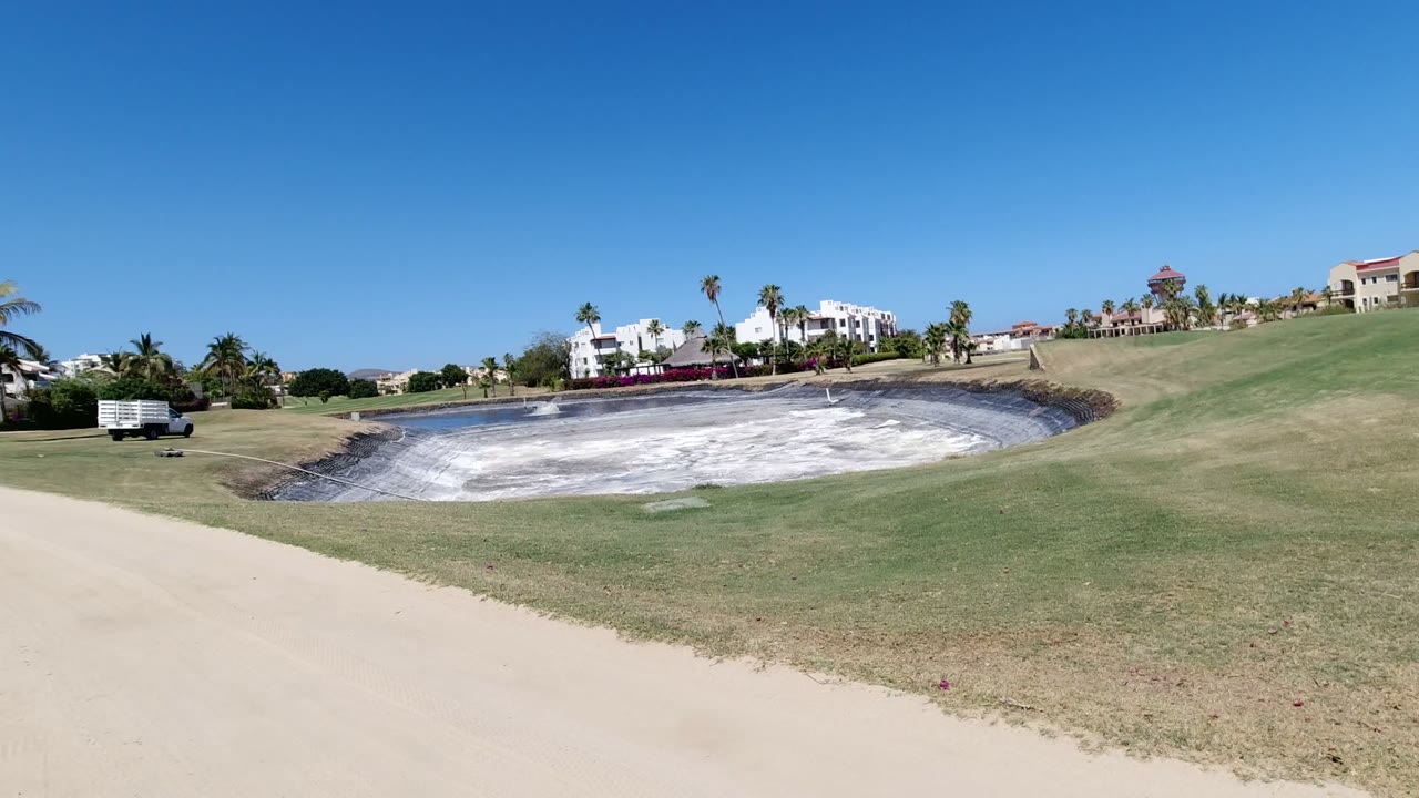 Los Cabos landscaped grounds with a curved sandy lane, white-lined water basin, palms, distant buildings, and no visible people.