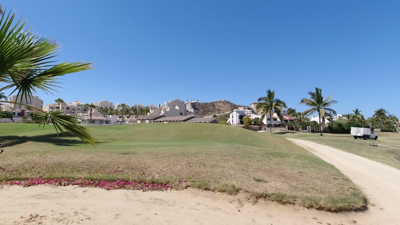 Opening frame of a Los Cabos, Mexico residential green space with palms, white buildings, a pale path, pink flowers in front, and no visible people.