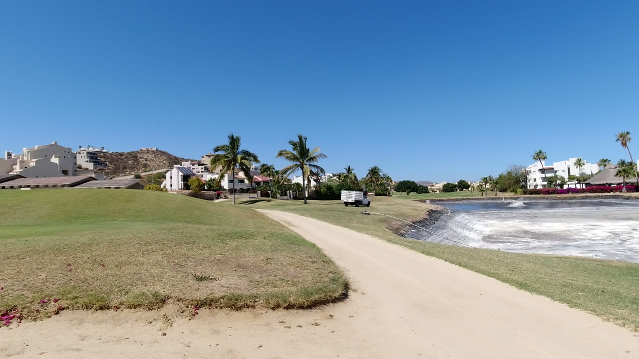 Wide resort lawn, path, palm trees, white buildings, and pale pond under blue sky.