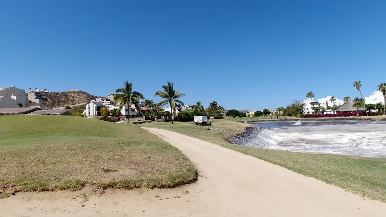 One second into the Los Cabos clip, the path and grass remain prominent while palm trees, white resort buildings, a pale pond, and no visible people fill the background.