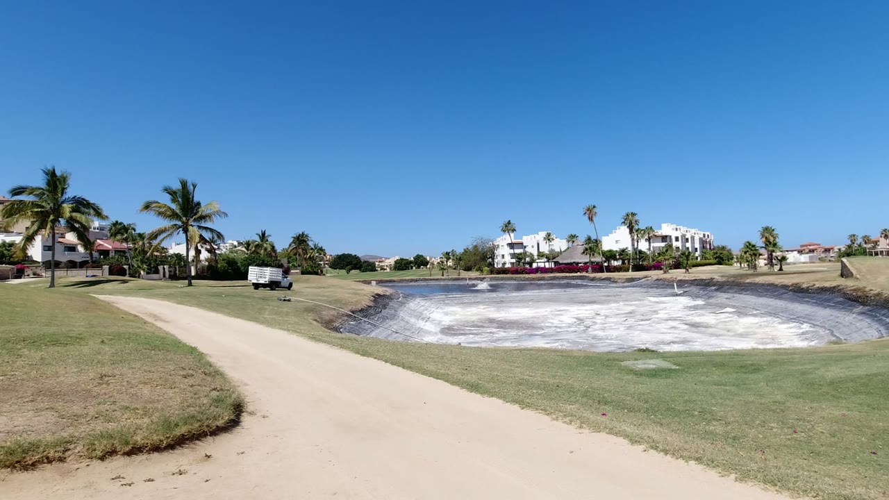 Golf course path beside a pale basin, green fairway, palms, and modern homes.