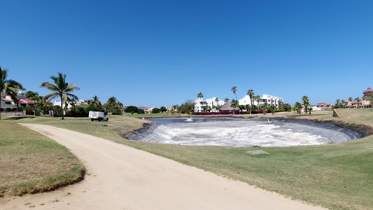 Wide Los Cabos fairway view showing the pale basin centered lower in frame, white homes across the background, palms along the grounds, and no visible people.