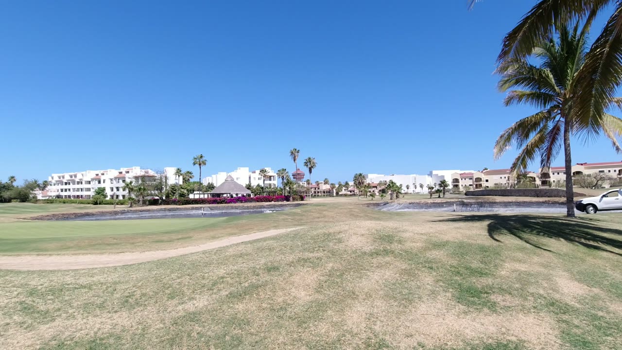 Opening frame of a Los Cabos grass landscape with a tall palm at the right edge, white low-rise buildings across the background, a water feature in the midground, and no visible people.