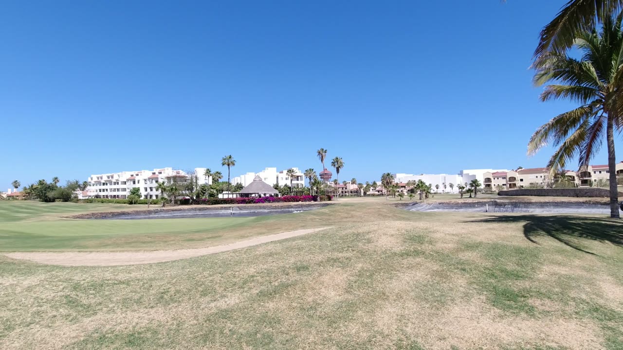 Los Cabos landscape view with a curved dirt path across dry grass, a pond or water feature to the left of center, white buildings in the background, scattered palms, and no visible people.