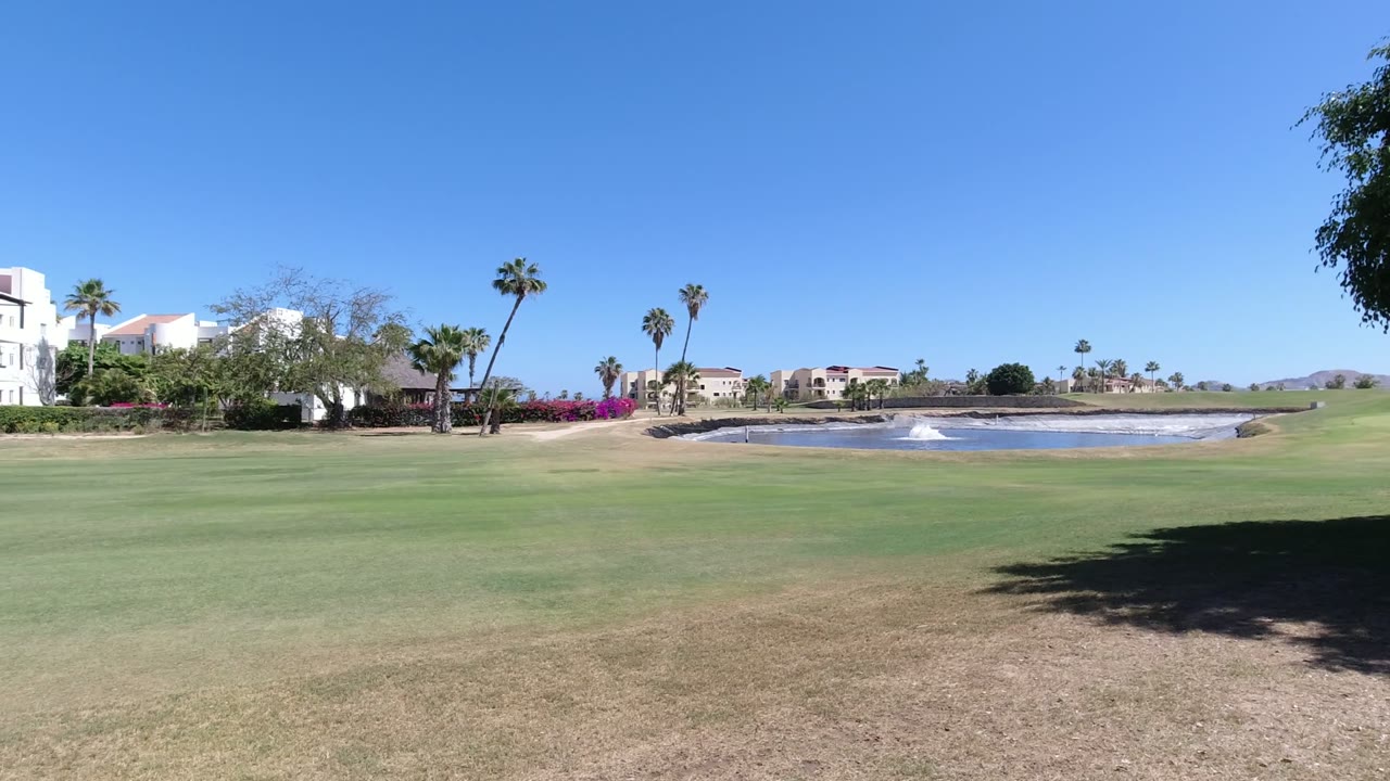 Wide Los Cabos golf course view with green and brown turf, white resort-style buildings at left, palm trees, a small water feature at right, blue sky, and no visible people.