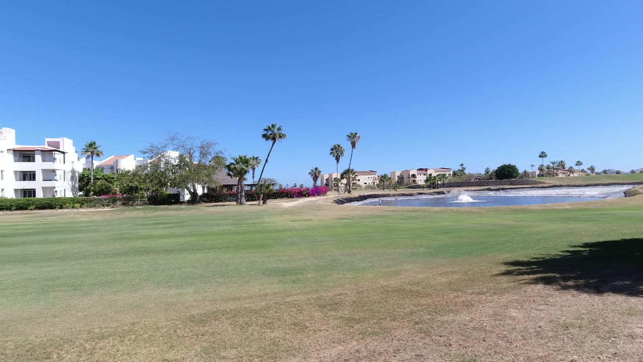 Los Cabos golf course frame with the lake more centered, white buildings on the left, palm trees across the horizon, bright blue sky, and no visible people.